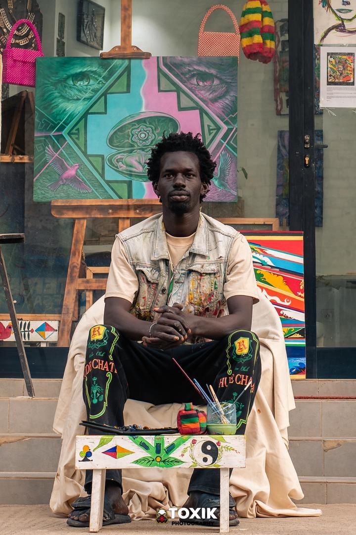 Nalla Thioye assis devant son atelier, veste tachée de peinture, une toile sur chevalet derrière lui